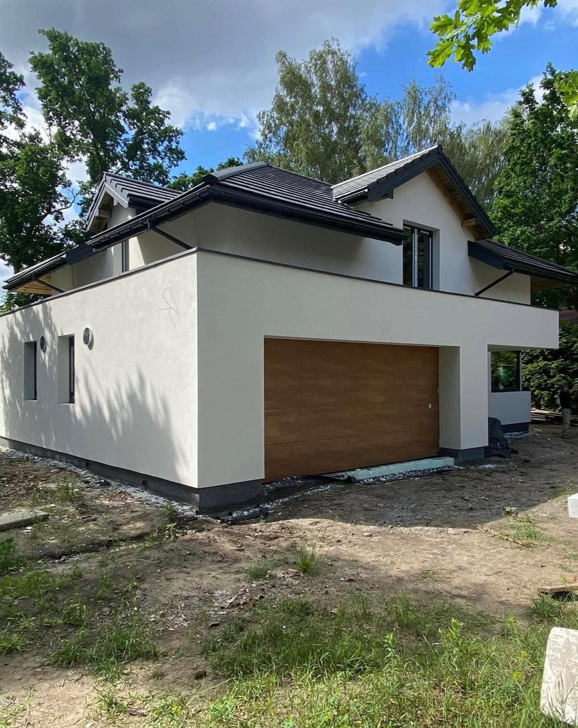 Nowoczesny, dwukondygnacyjny dom z jasną elewacją i brązową bramą garażową, otoczony zielenią i błękitnym niebem.