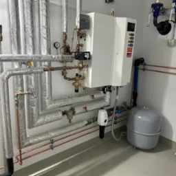 Złożona instalacja grzewcza z rurami w srebrnej izolacji, kotłem gazowym z panelem sterowania, zbiornikiem wyr&oacute;wnawczym oraz fragmentem umywalki w rogu pomieszczenia.