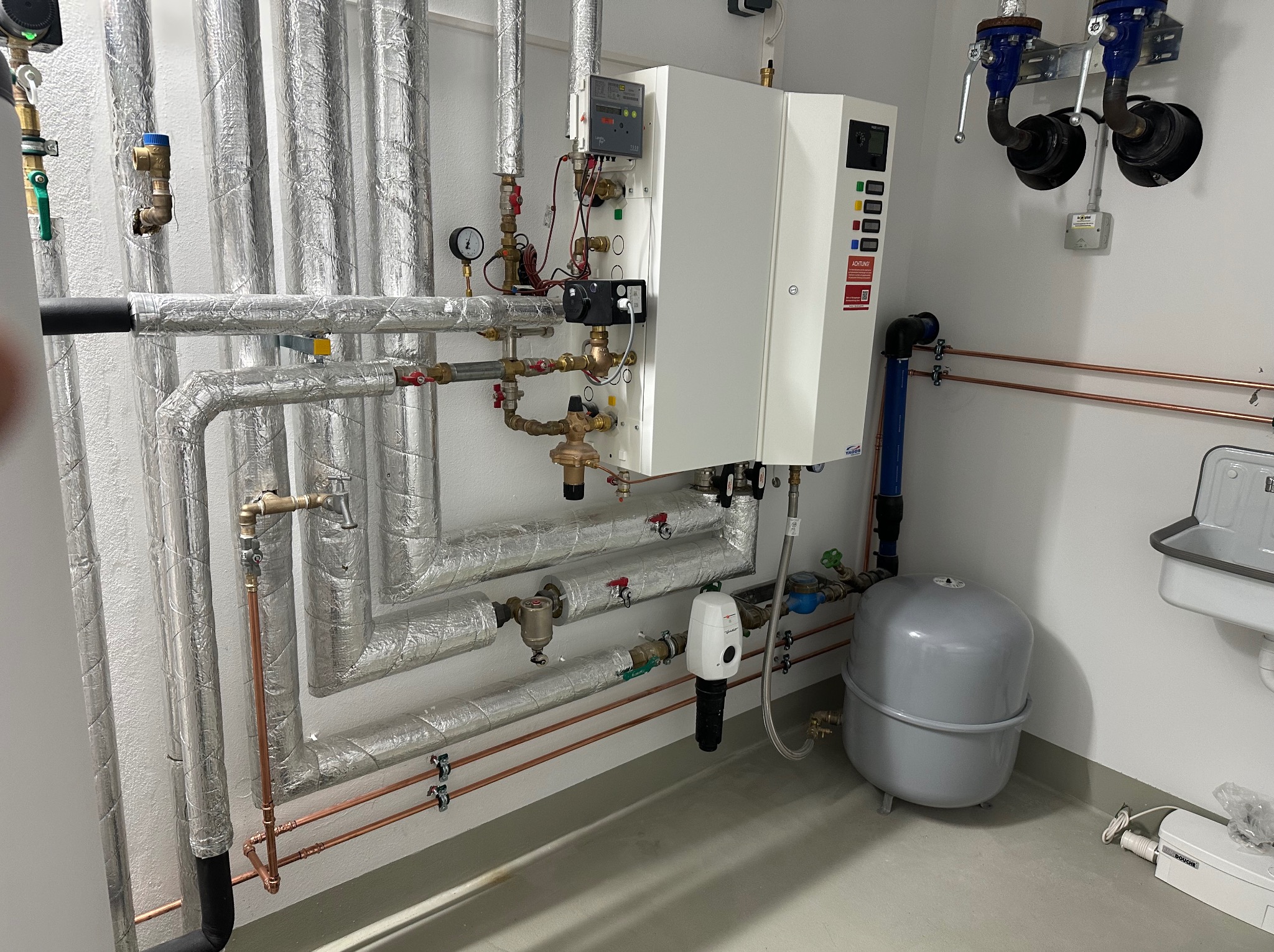 Złożona instalacja grzewcza z rurami w srebrnej izolacji, kotłem gazowym z panelem sterowania, zbiornikiem wyrównawczym oraz fragmentem umywalki w rogu pomieszczenia.