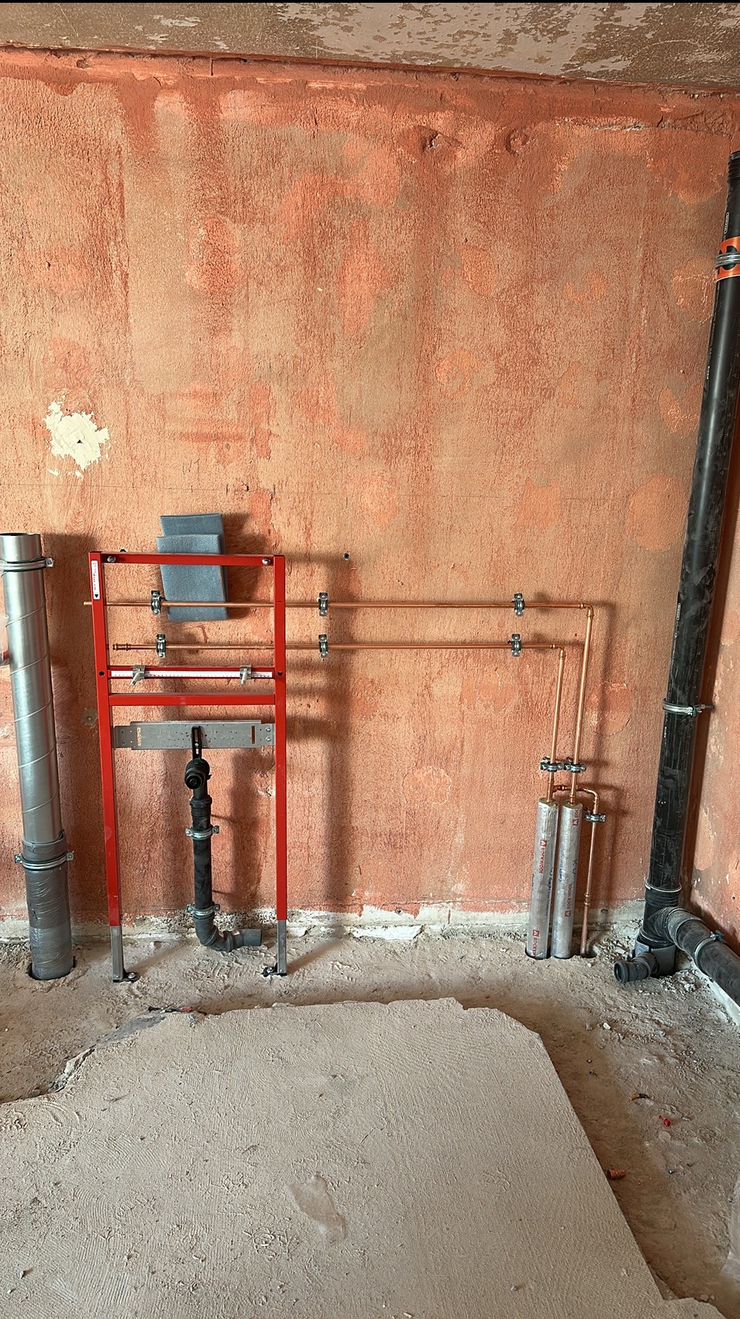 Surowa ściana w odcieniach pomarańczy z widocznymi elementami instalacji hydraulicznej: stelaż podtynkowy z rurami miedzianymi, pionowy odpływ kanalizacyjny i pionowa rura wentylacyjna, wszystko...