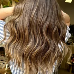 Widok z tyłu na długie, falowane włosy z koloryzacją typu sombre, przechodzące od ciemnego brązu u nasady do jasnego blondu na końcach. Modelka ubrana w sukienkę w biało-niebieskie paski.