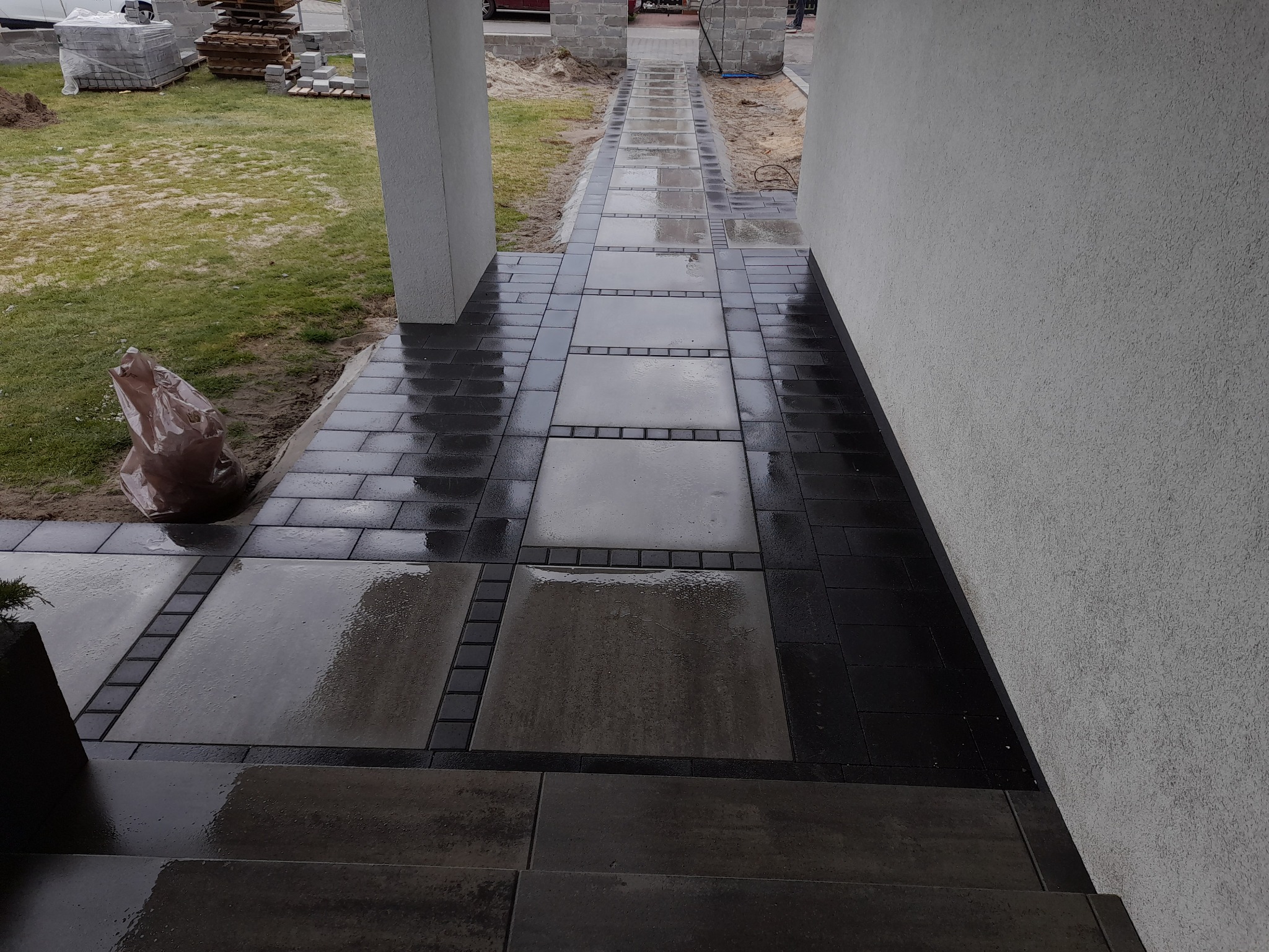 Nowo ułożona, mokra nawierzchnia z kostki brukowej w odcieniach szarości i czerni prowadząca do wejścia domu, z widocznym workiem budowlanym i paletami kostki w tle.