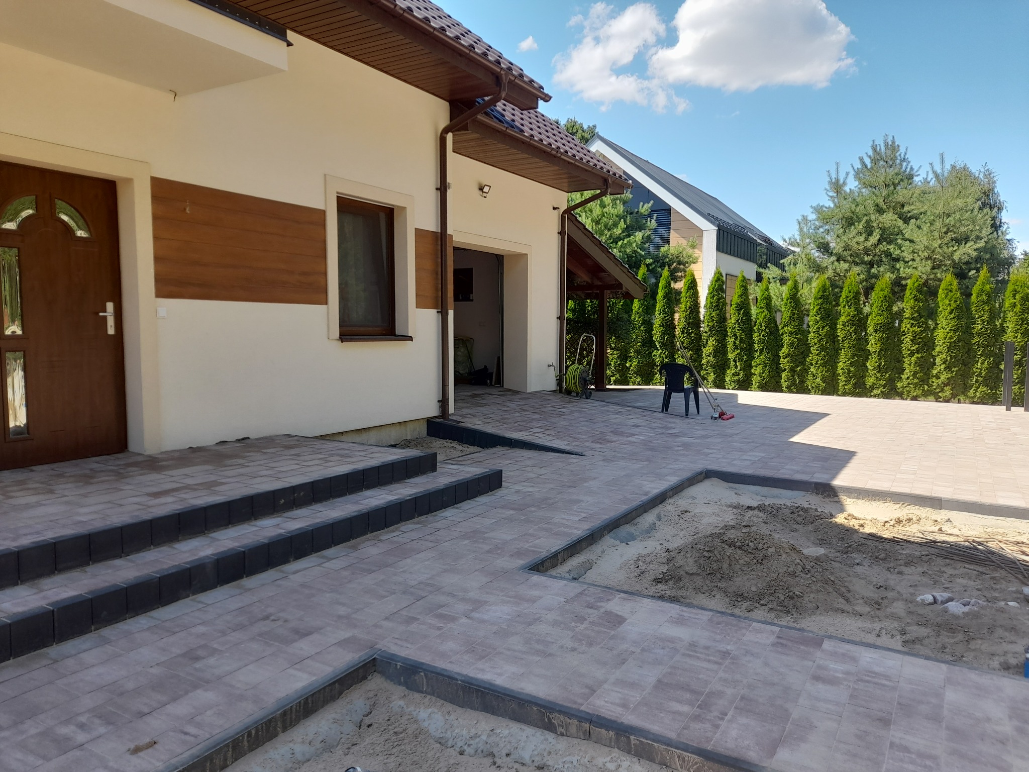 Nowo ułożona kostka brukowa w odcieniach szarości prowadząca do domu z brązowymi drzwiami i drewnianą elewacją, widoczny fragment przygotowywanej rabaty z ziemią.