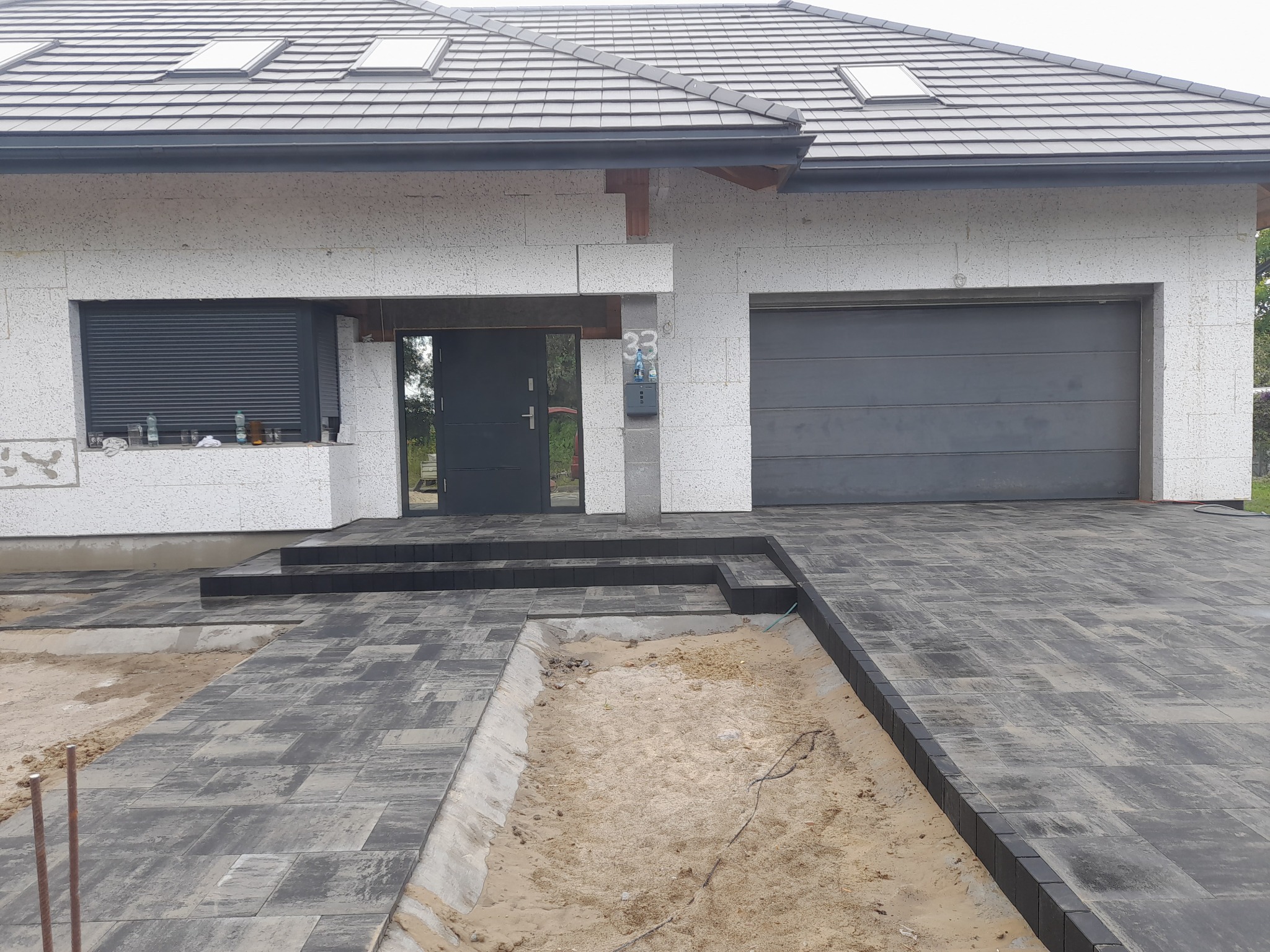 Układanie kostki brukowej w kolorze grafitowym przed nowoczesnym domem z białego kamienia, widoczny fragment wykopu pod dalszą część nawierzchni.