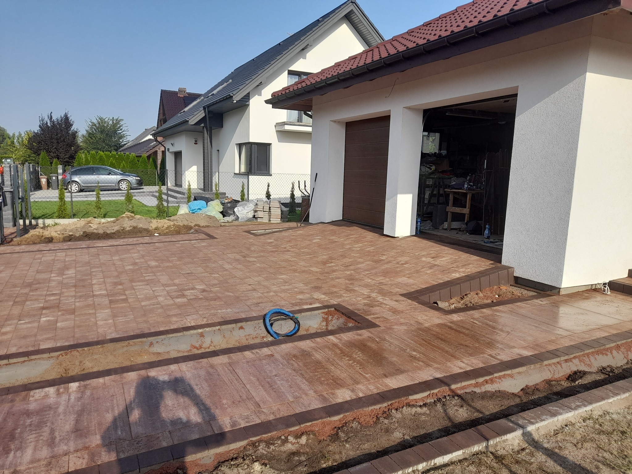 Częściowo ułożona, czerwono-brązowa nawierzchnia z kostki brukowej przed domem z garażem; widoczne przygotowane koryta pod instalacje i obrzeża.