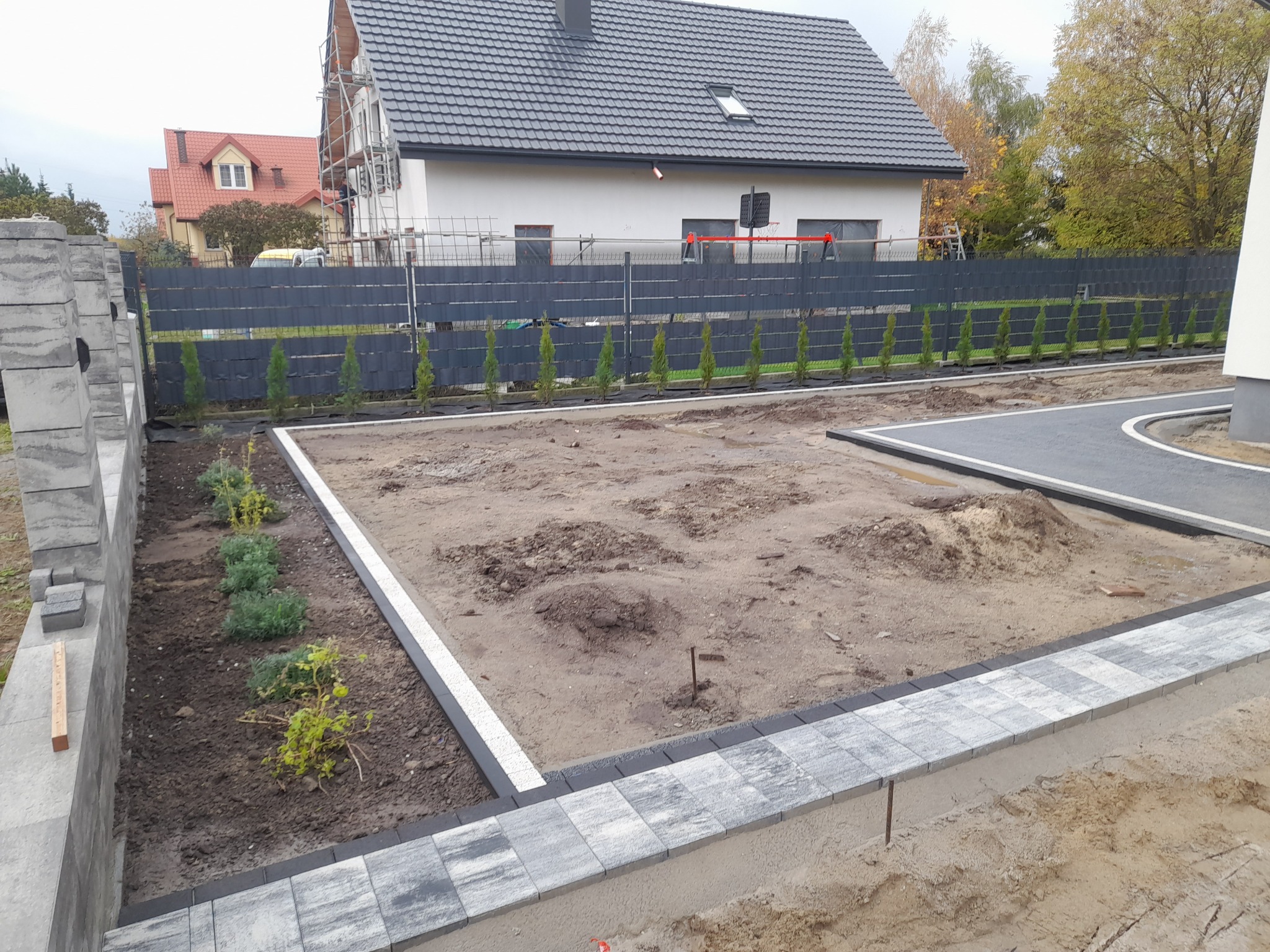 Przygotowany teren pod trawnik z obrzeżami z kostki granitowej i w tle rząd nowo posadzonych tuj.