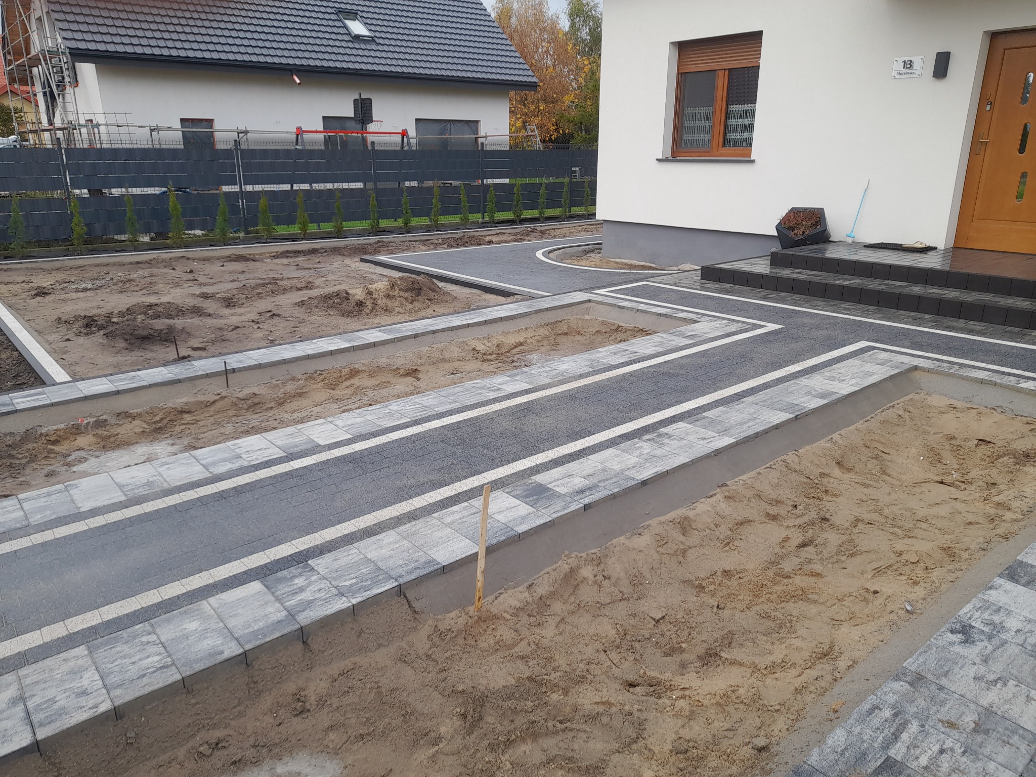 Fragment posesji z częściowo ułożoną kostką brukową w odcieniach szarości i bieli, widoczne przygotowane podłoże pod dalsze prace, dom w tle.