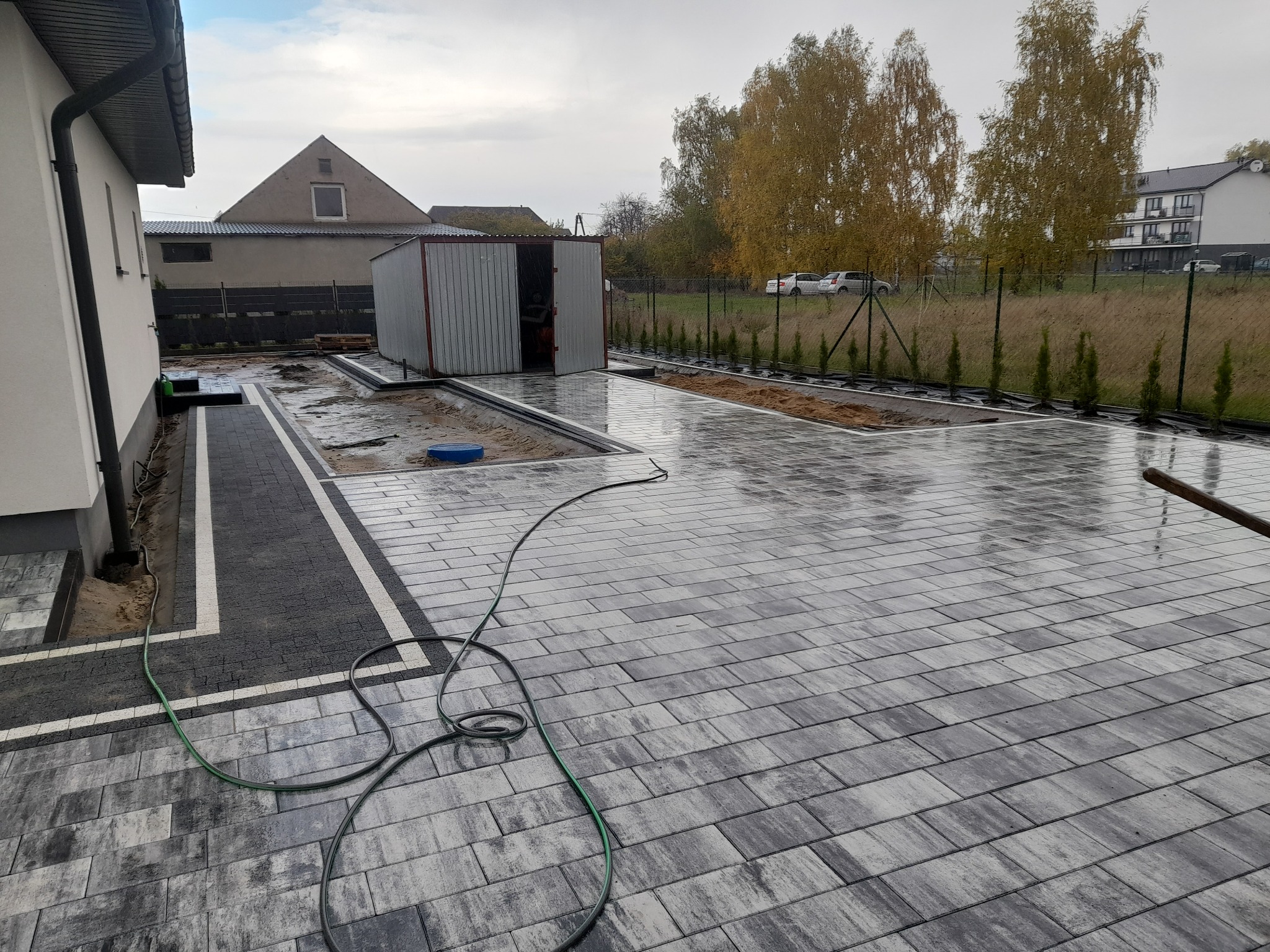 Ułożona, wilgotna kostka brukowa w odcieniach szarości i czerni z białymi obramowaniami, widoczny fragment domu, metalowy garaż i zielony wąż ogrodowy.
