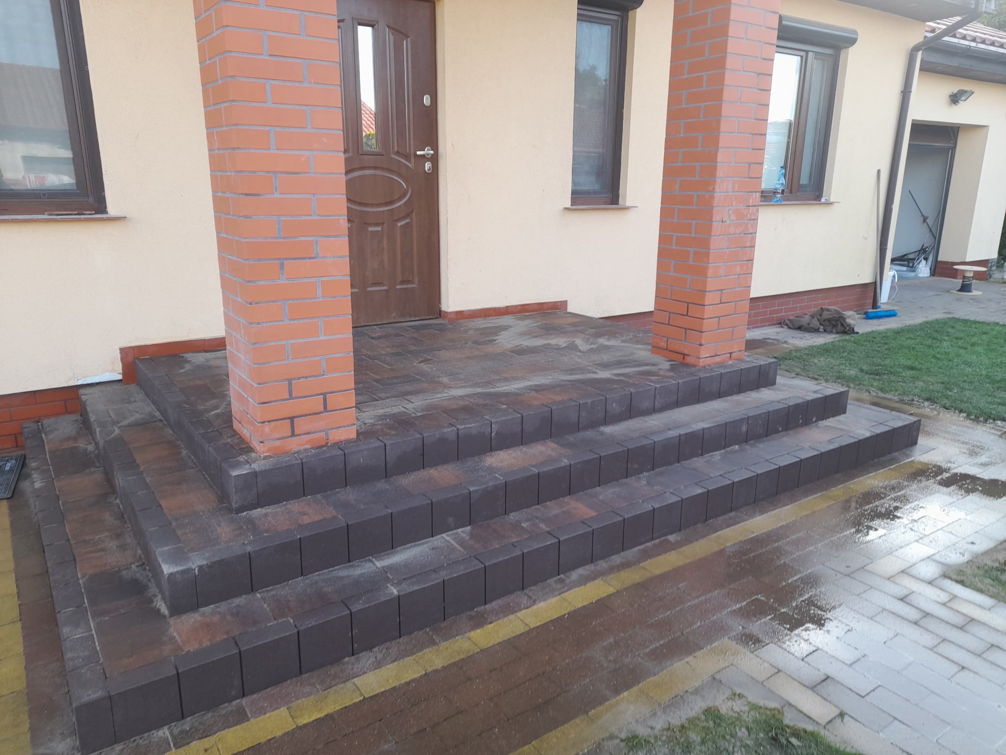 Schody wejściowe do domu wyłożone kostką w odcieniach brązu i szarości, z kolumnami z cegły po bokach, na mokrym podłożu.