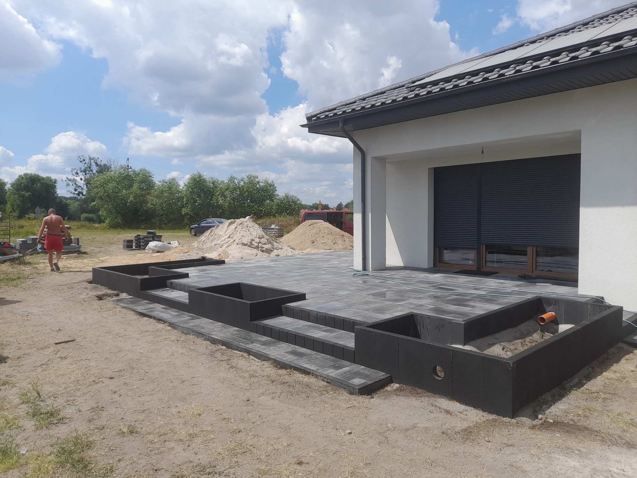 Układanie szarej kostki brukowej na tarasie z donicami, widoczny fragment domu z szarymi roletami, piasek i pracownik w tle.