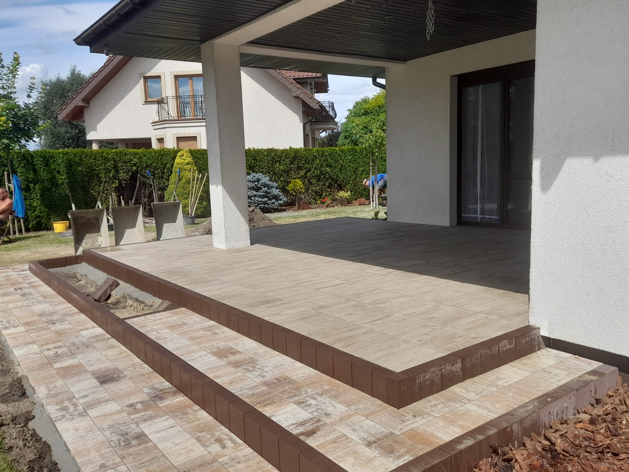 Wyłożony płytkami taras z dwoma stopniami i obrzeżem z kostki w kolorze brązowym, widok na dom i zadbany ogród.