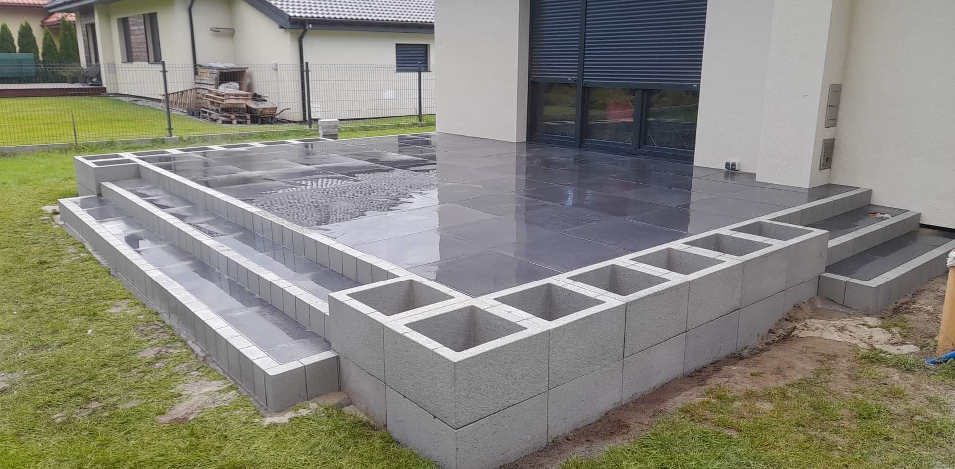 Nowoczesny taras z ciemnej, błyszczącej kostki, obrzeża z szarego betonu, trzy stopnie wejściowe, widoczne odbicie nieba na mokrej powierzchni.