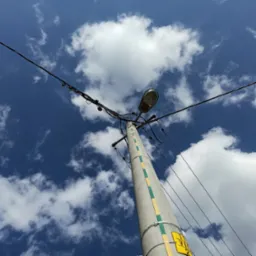 Betonowy słup energetyczny z lampą uliczną i plątaniną kabli na tle błękitnego nieba z białymi obłokami, widok z dołu.