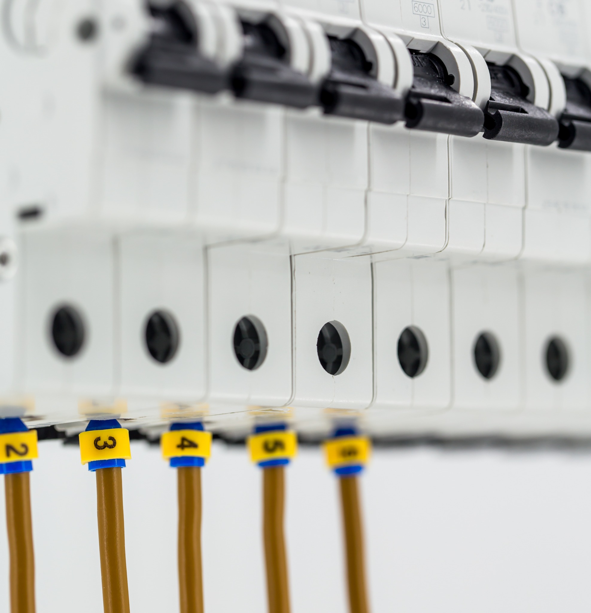 Szczegółowe ujęcie panelu elektrycznego z wyłącznikami, widoczne brązowe przewody z numerowanymi, żółto-niebieskimi oznaczeniami (2, 3, 4, 5, 6), na białym tle.