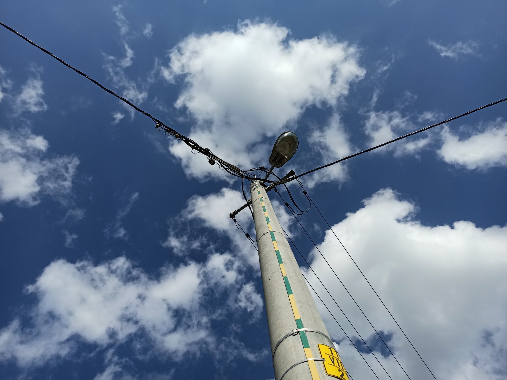 Betonowy słup energetyczny z lampą uliczną i plątaniną kabli na tle błękitnego nieba z białymi obłokami, widok z dołu.