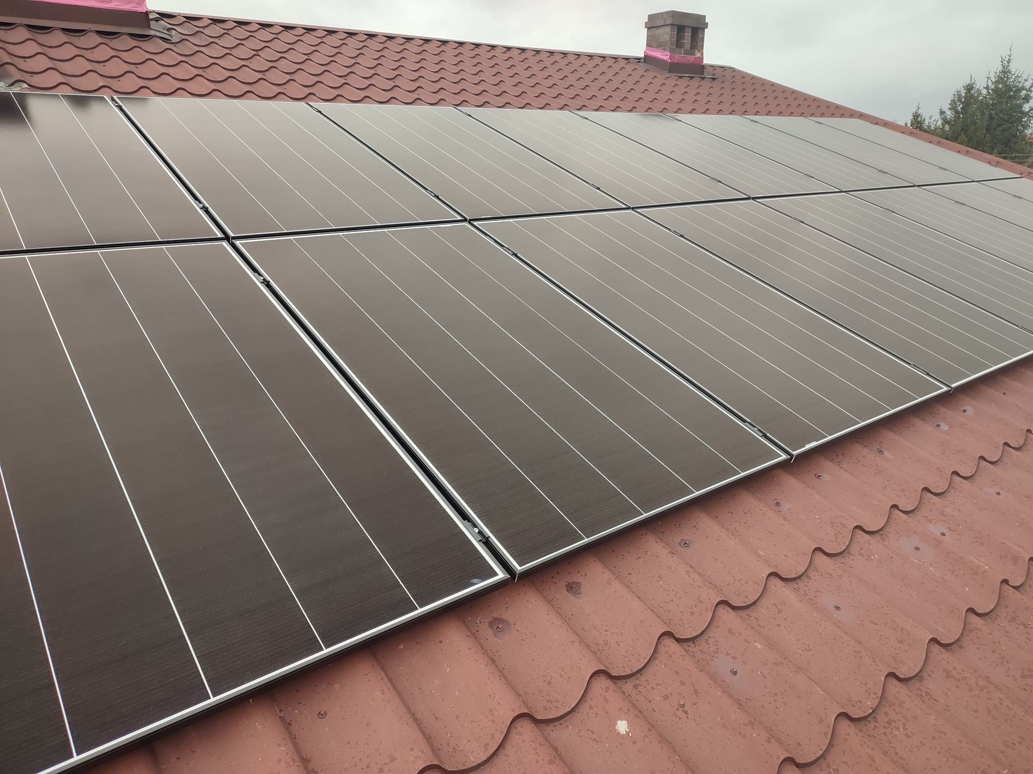 Panele słoneczne zamontowane na czerwonym dachu z blachodachówki, widoczny komin z różowym zabezpieczeniem.