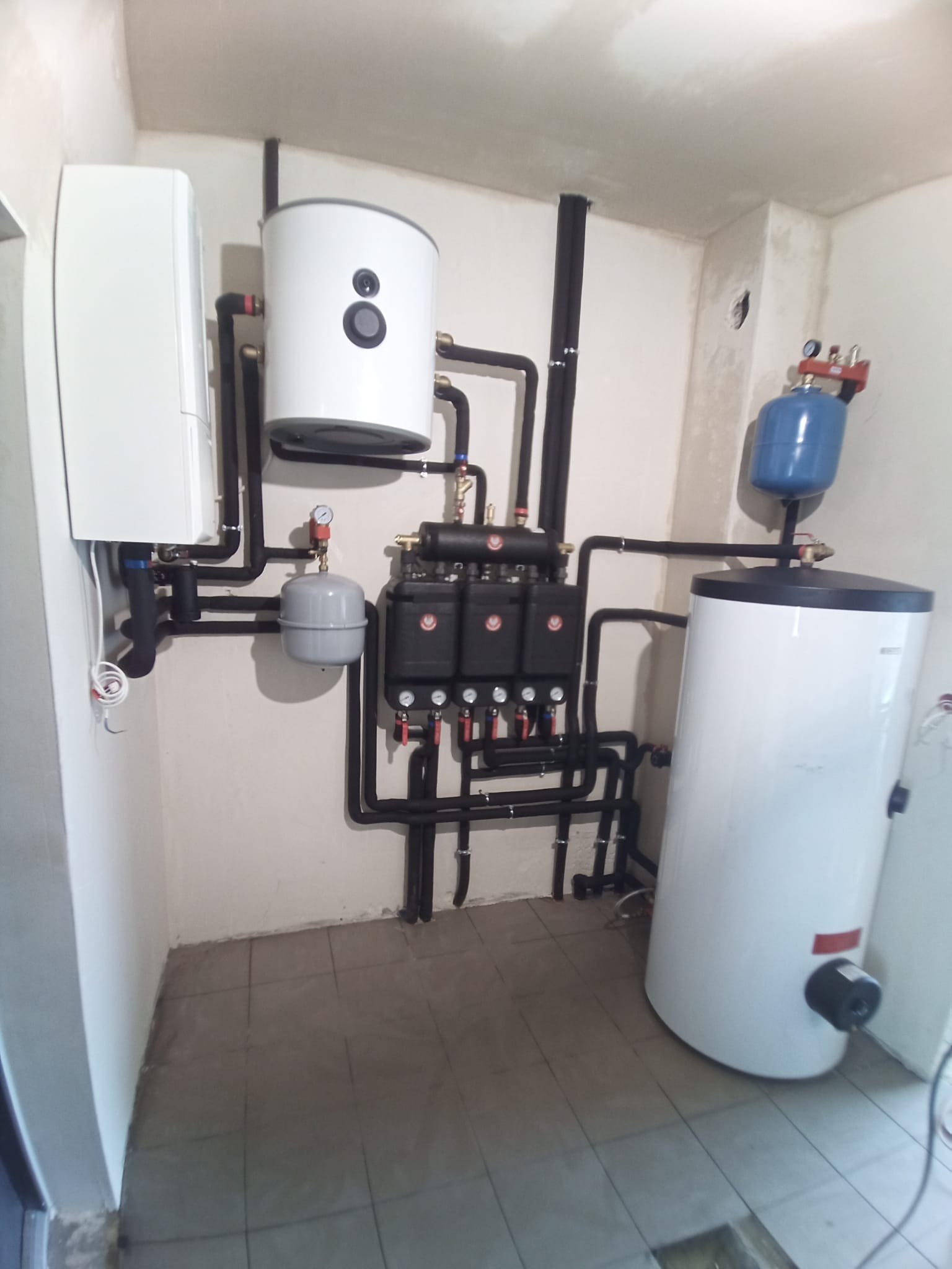 Instalacja grzewcza w kotłowni: białe zasobniki ciepłej wody, czarne rury, szara armatura, manometry z czerwonymi wskazówkami.