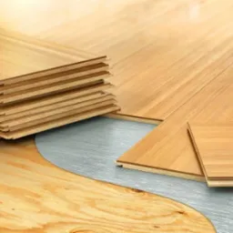 Stos jasnych paneli podłogowych ułożony na srebrnej folii izolacyjnej, widoczna struktura drewna i warstwy paneli.