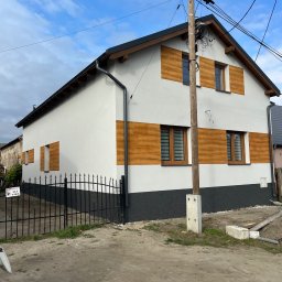 Delta Home - Wykończona elewacja domu z poziomymi panelami imitującymi drewno i świeżo pomalowaną biało-szarą fasadą. Widoczny słup energetyczny i fragment ogrodzenia.