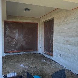 Delta Home - Zadaszony taras w trakcie wykończenia: ściany z szarymi panelami imitującymi drewno, okna zabezpieczone folią, podłoże przygotowane do dalszych prac.