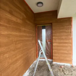 Elewacja budynku wykończona w stylu imitującym drewno, z drabiną opartą o osłonięte drzwi, detale wykończeniowe w kolorze pomarańczowym.