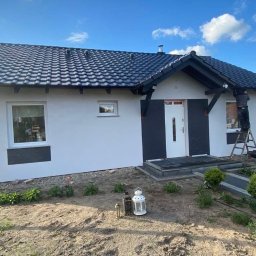 Delta Home - Biała elewacja domu z czarnymi akcentami, pracownik na drabinie przy okapie. Nowe okna, dachówka i zagospodarowany teren wokół domu.