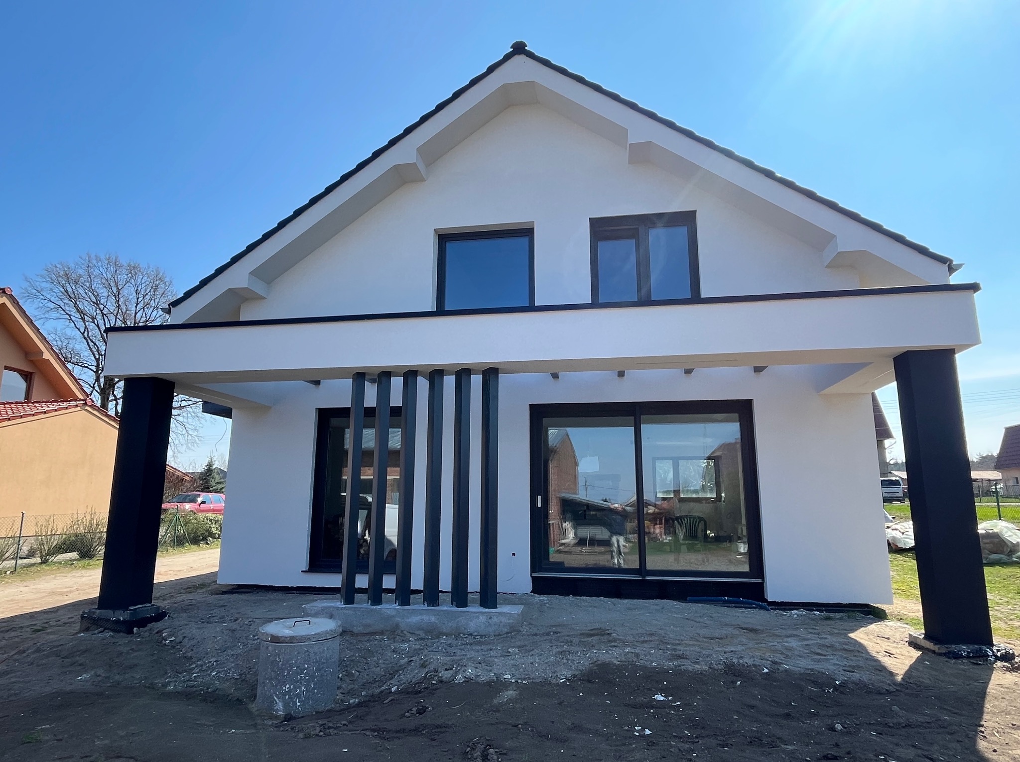 Biały dom jednorodzinny z czarnymi oknami i kolumnami werandy w słoneczny dzień. Widoczne detale elewacji i otoczenie budynku.