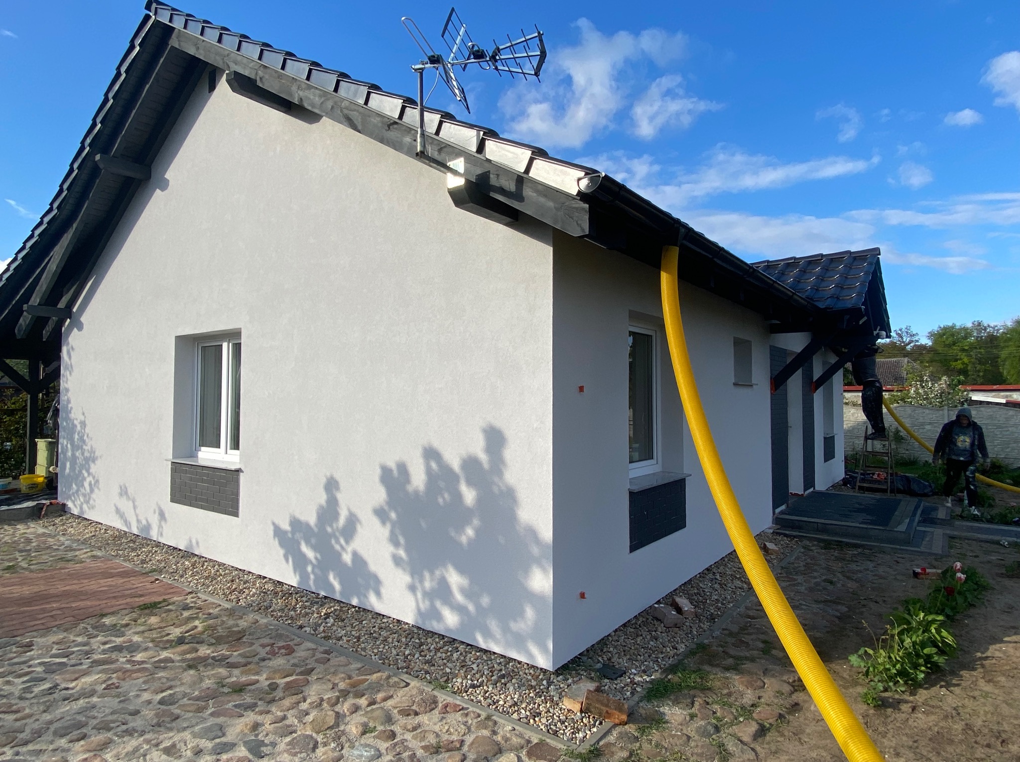Biały dom z ciemną dachówką po ociepleniu elewacji, widoczny żółty wąż do natrysku. Na dachu antena. Kostka brukowa wokół budynku. Błękitne niebo.