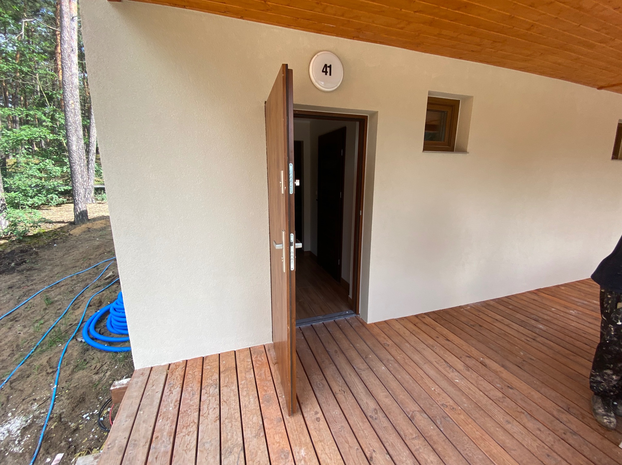 Elewacja budynku z numerem 41, drewniany taras, otwarte drzwi. Ściana w jasnym odcieniu, drewniana podbitka dachowa. Widok na las w tle.