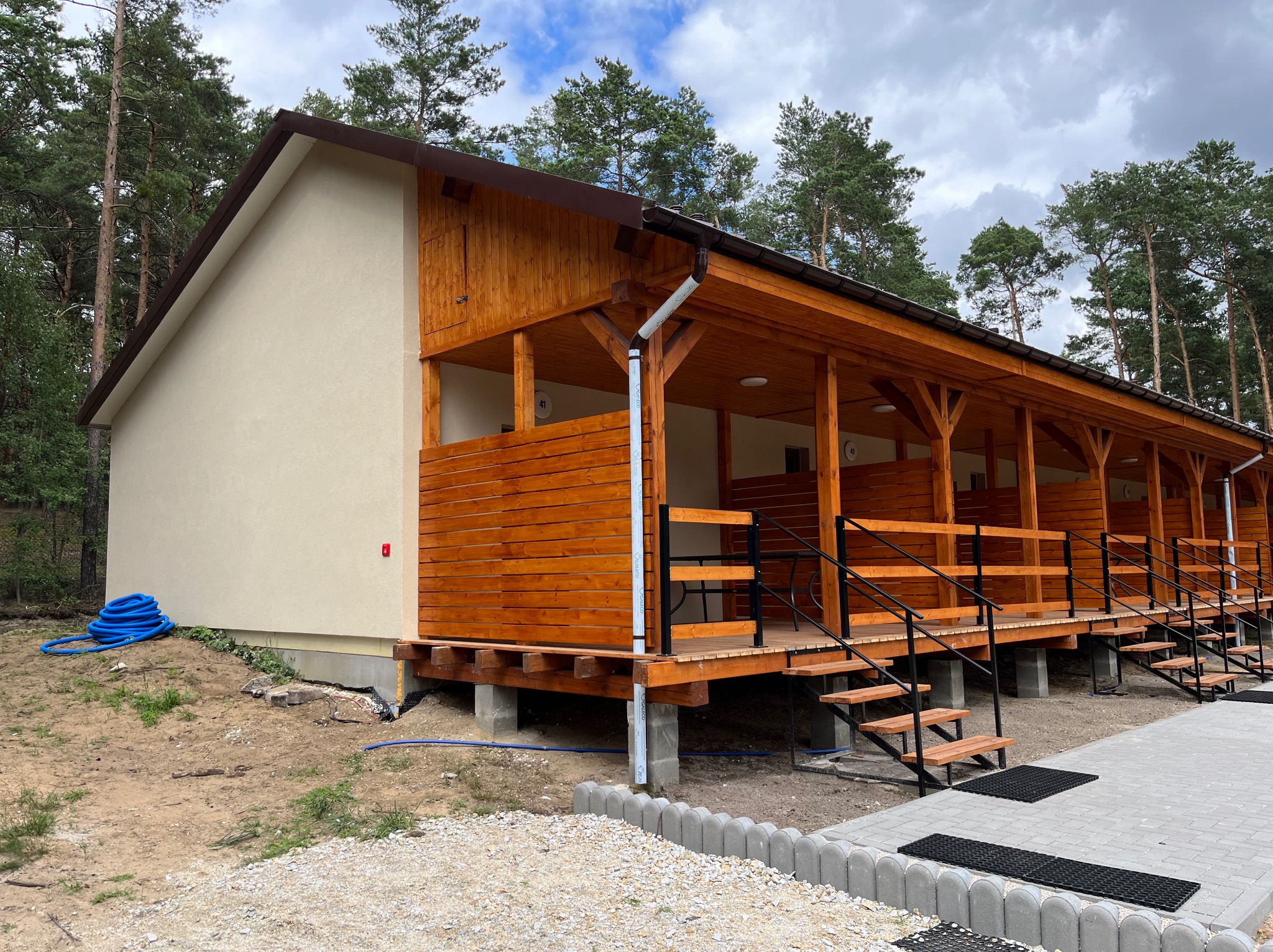 Elewacja parterowego budynku z drewnianą werandą i schodami. Jasna fasada kontrastuje z brązowym drewnem. W tle widoczne drzewa.