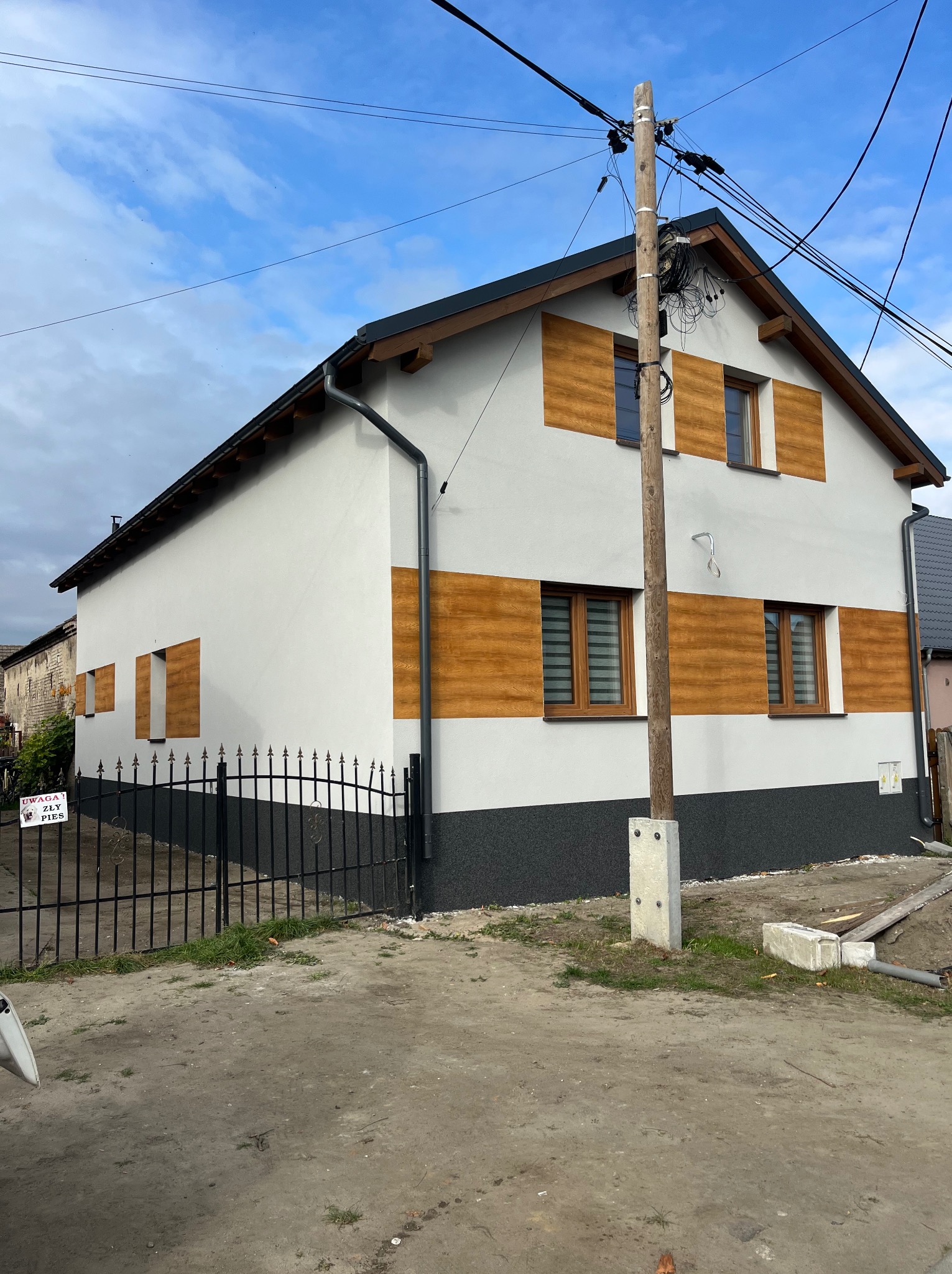 Wykończona elewacja domu z poziomymi panelami imitującymi drewno i świeżo pomalowaną biało-szarą fasadą. Widoczny słup energetyczny i fragment ogrodzenia.