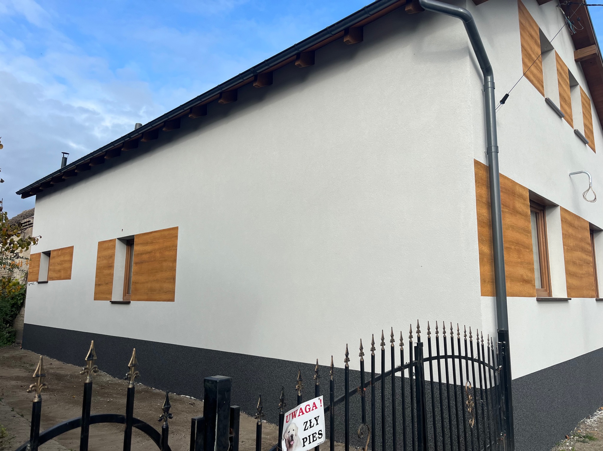 Biała elewacja domu z drewnopodobnymi panelami okiennymi i ciemną podmurówką, widoczna zza czarnego, metalowego ogrodzenia z tabliczką 'Uwaga! Zły pies'.