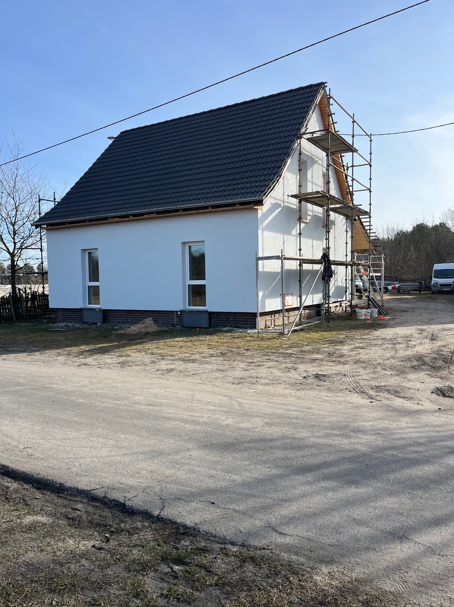 Biały dom z czarnym dachem w trakcie prac elewacyjnych, otoczony rusztowaniami. Widoczne okna i elementy wykończeniowe. Częściowo otynkowana ściana.