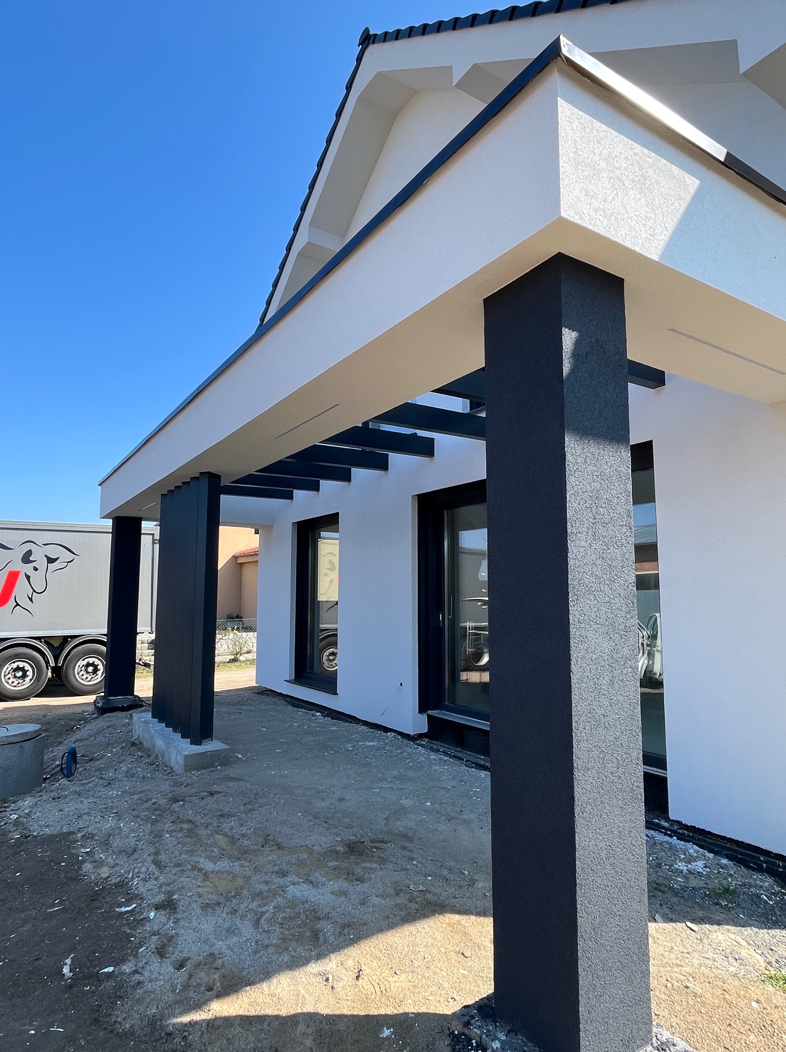Nowoczesny dom z elewacją w kolorze białym i antracytowymi kolumnami. Widoczny fragment elewacji i zarys ciężarówki w tle. Słoneczny dzień.