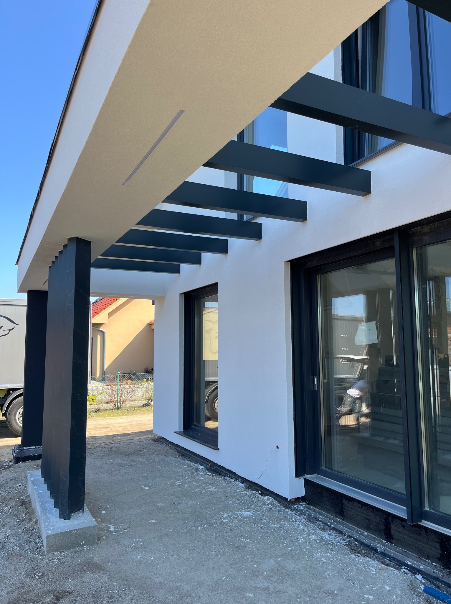 Nowoczesna elewacja domu z minimalistycznym zadaszeniem i dużymi oknami. Widoczne detale wykończenia, jasna kolorystyka i elementy konstrukcyjne.