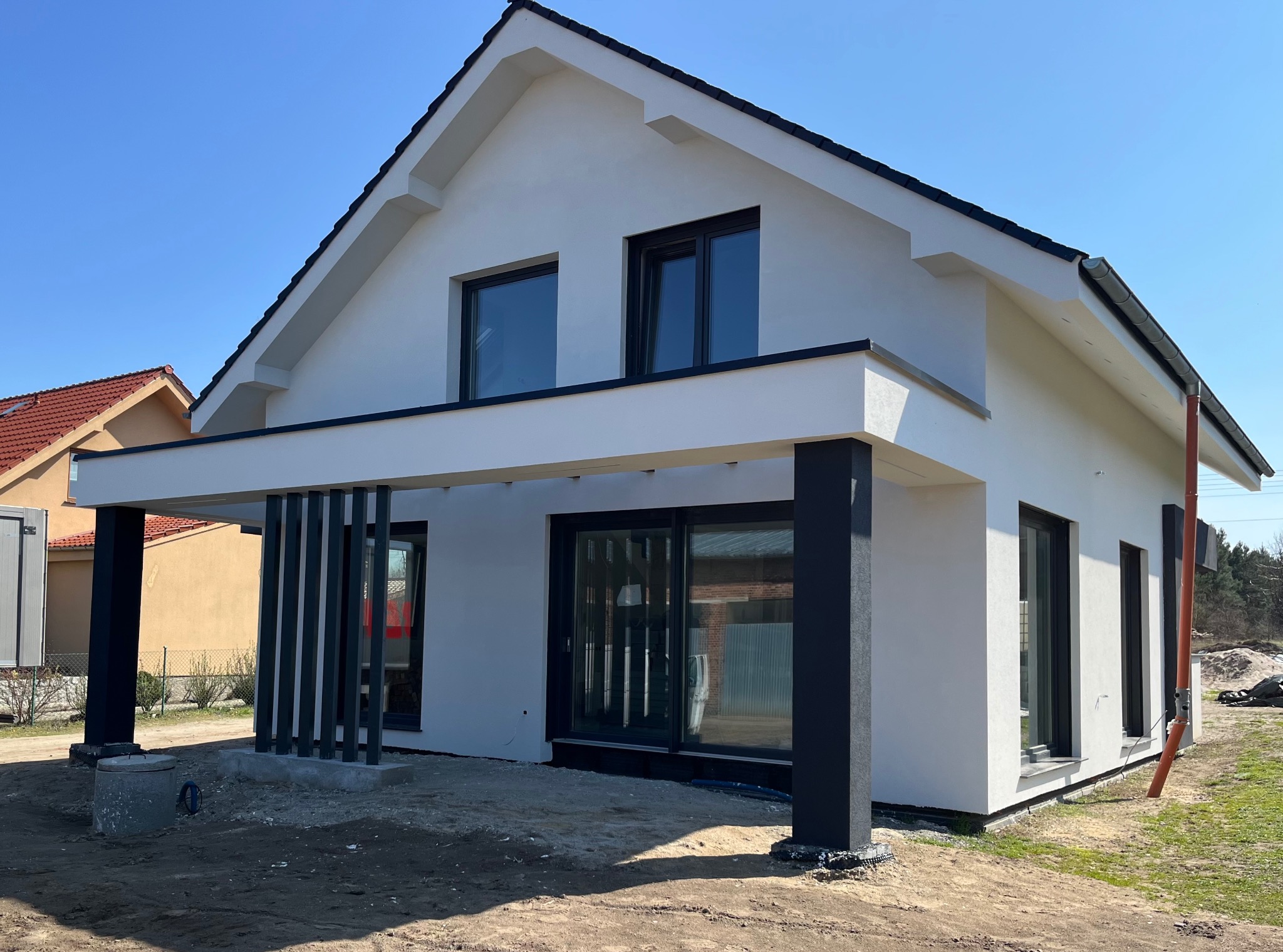 Biały dom z czarnym dachem i stolarką okienną w słoneczny dzień. Widoczne elementy elewacji, orynnowanie i fragment terenu przed budynkiem.