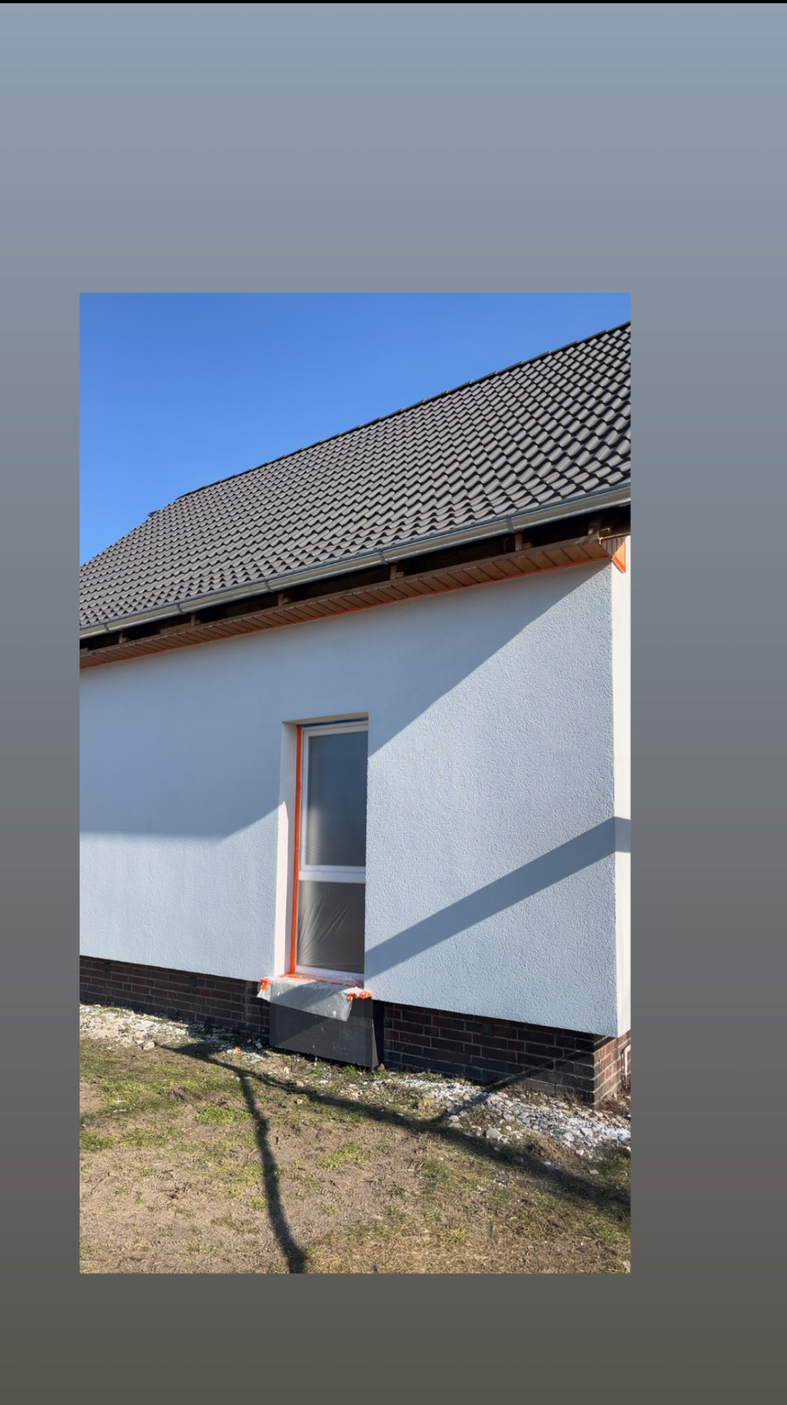 Boczny widok domu z białą elewacją i szarym dachem. Okno zabezpieczone folią, widoczne ceglane fundamenty i fragment trawnika. Słoneczny dzień, błękitne niebo.