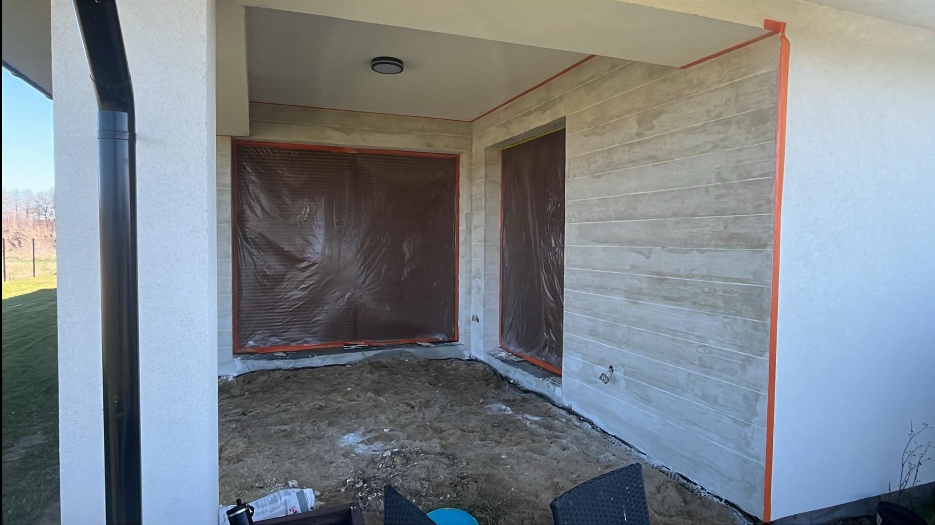 Zadaszony taras w trakcie wykończenia: ściany z szarymi panelami imitującymi drewno, okna zabezpieczone folią, podłoże przygotowane do dalszych prac.