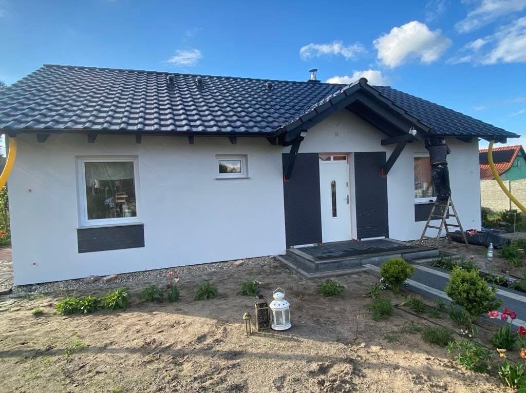 Biała elewacja domu z czarnymi akcentami, pracownik na drabinie przy okapie. Nowe okna, dachówka i zagospodarowany teren wokół domu.