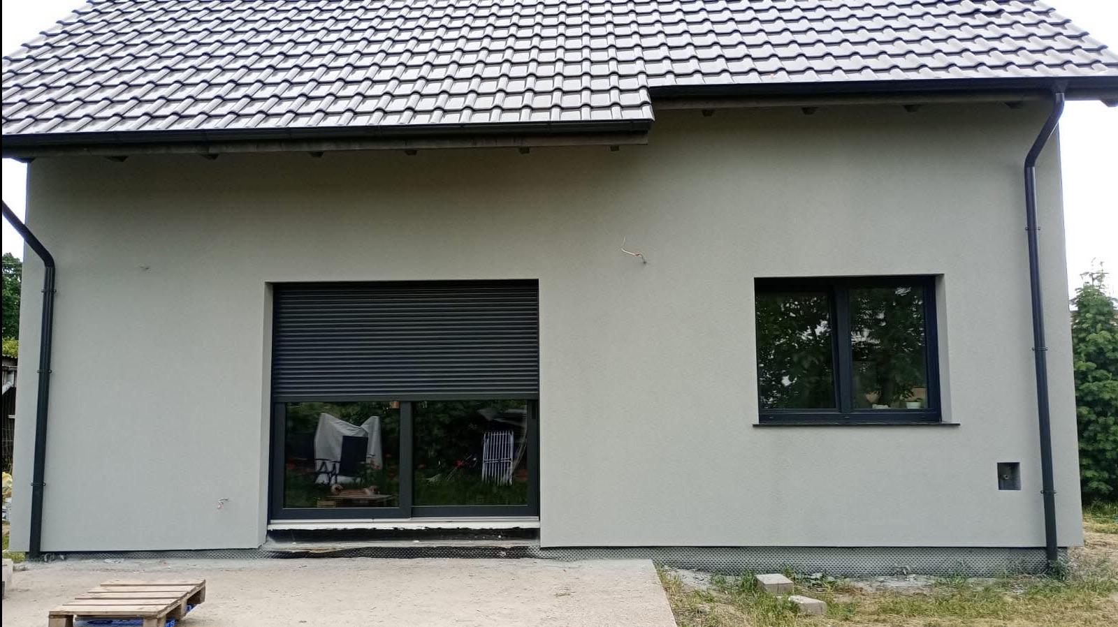 Szara elewacja domu jednorodzinnego z ciemnymi oknami i roletą zewnętrzną. Widoczny fragment dachu z czarnej dachówki i rynna. Ujęcie z poziomu gruntu.