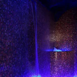 Wnętrze z mozaiką w odcieniach czerwieni i brązu, podświetlone intensywnym niebieskim światłem, z kaskadą wody spływającą po ścianie do małego basenu z białymi kamieniami.