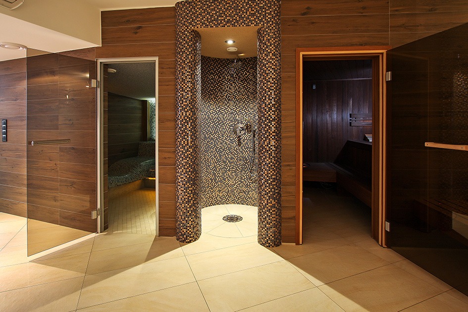 Wnętrze z mozaikową kabiną prysznicową między wejściami do strefy relaksu i sauny, drewniane wykończenia ścian, jasna podłoga z płytek.