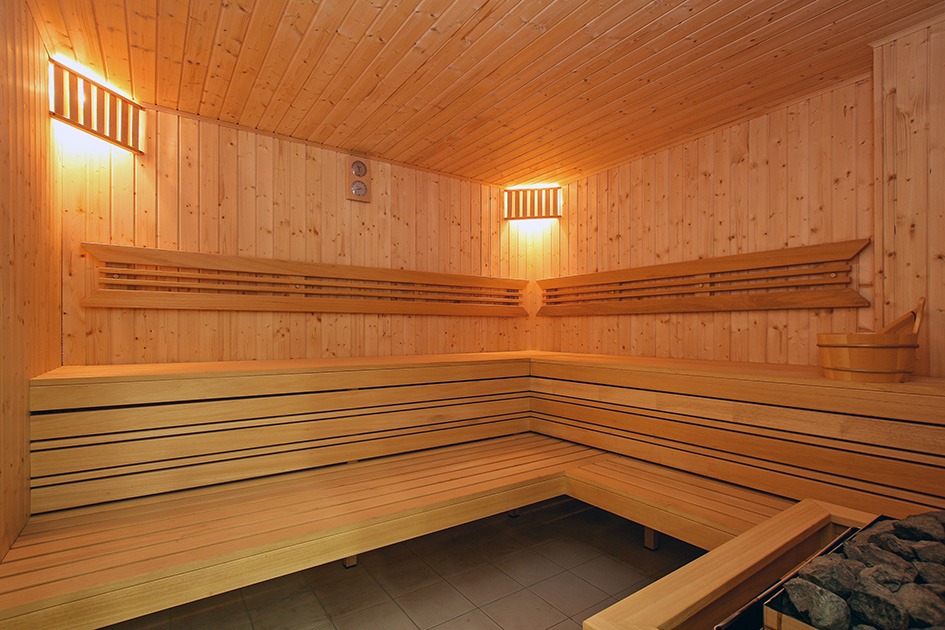 Wnętrze sauny wykończone drewnem, z ławkami na różnych poziomach, oświetleniem punktowym i widocznym wiaderkiem oraz kamieniami grzewczymi.