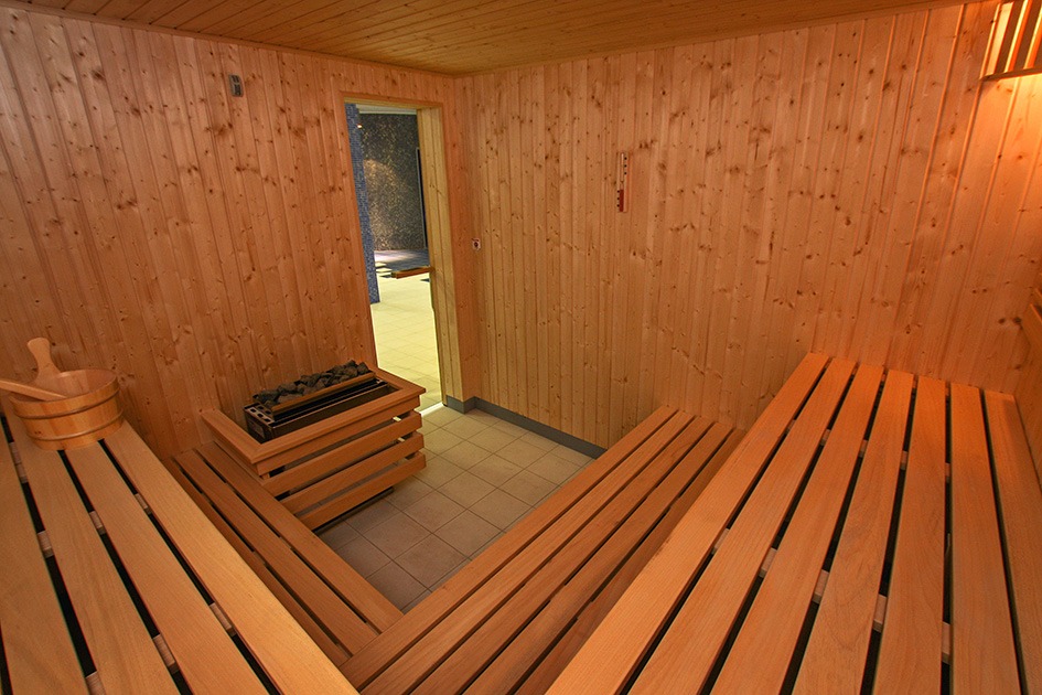 Wnętrze wykończonej drewnem sauny z ławkami, piecem na kamienie i akcesoriami, widok na przyległe pomieszczenie z mozaiką na ścianie.