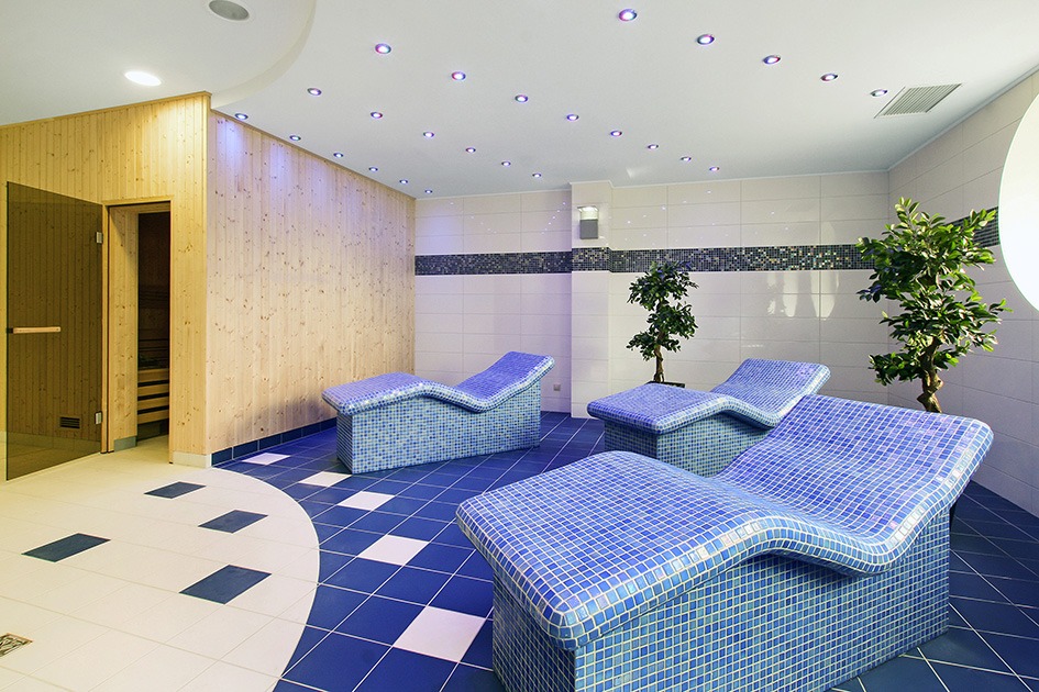 Relaksacyjna strefa sauny z trzema mozaikowymi leżankami w odcieniach błękitu, drzewkami w donicach i wejściem do sauny wykończonym drewnem.