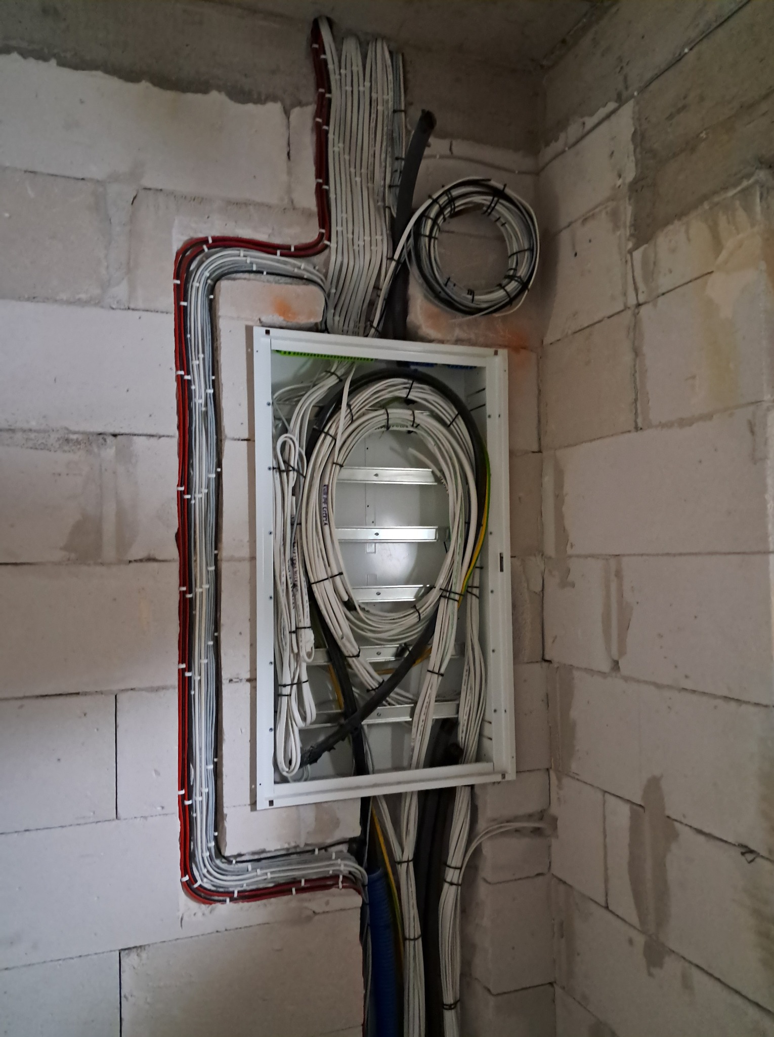 Rozdzielnica elektryczna z poprowadzonymi przewodami w bruzdach ściany z betonu komórkowego. Kable ułożone w pętle. Przewody w kolorze białym, czarnym i czerwonym.