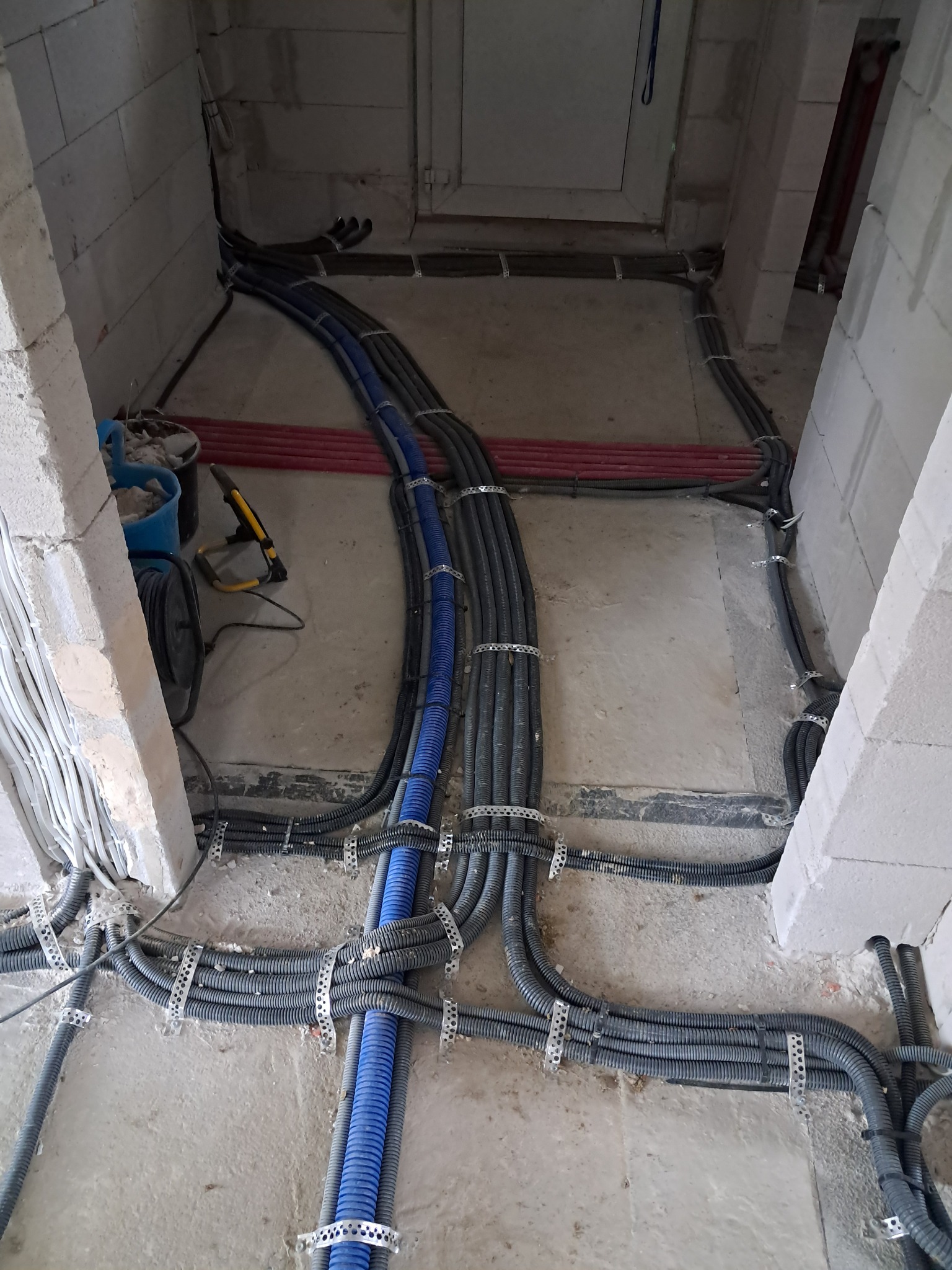 Kable w peszlach, częściowo niebieskich i szarych, ułożone na podłodze w surowym stanie, przymocowane metalowymi uchwytami do podłoża i ścian z betonu komórkowego.