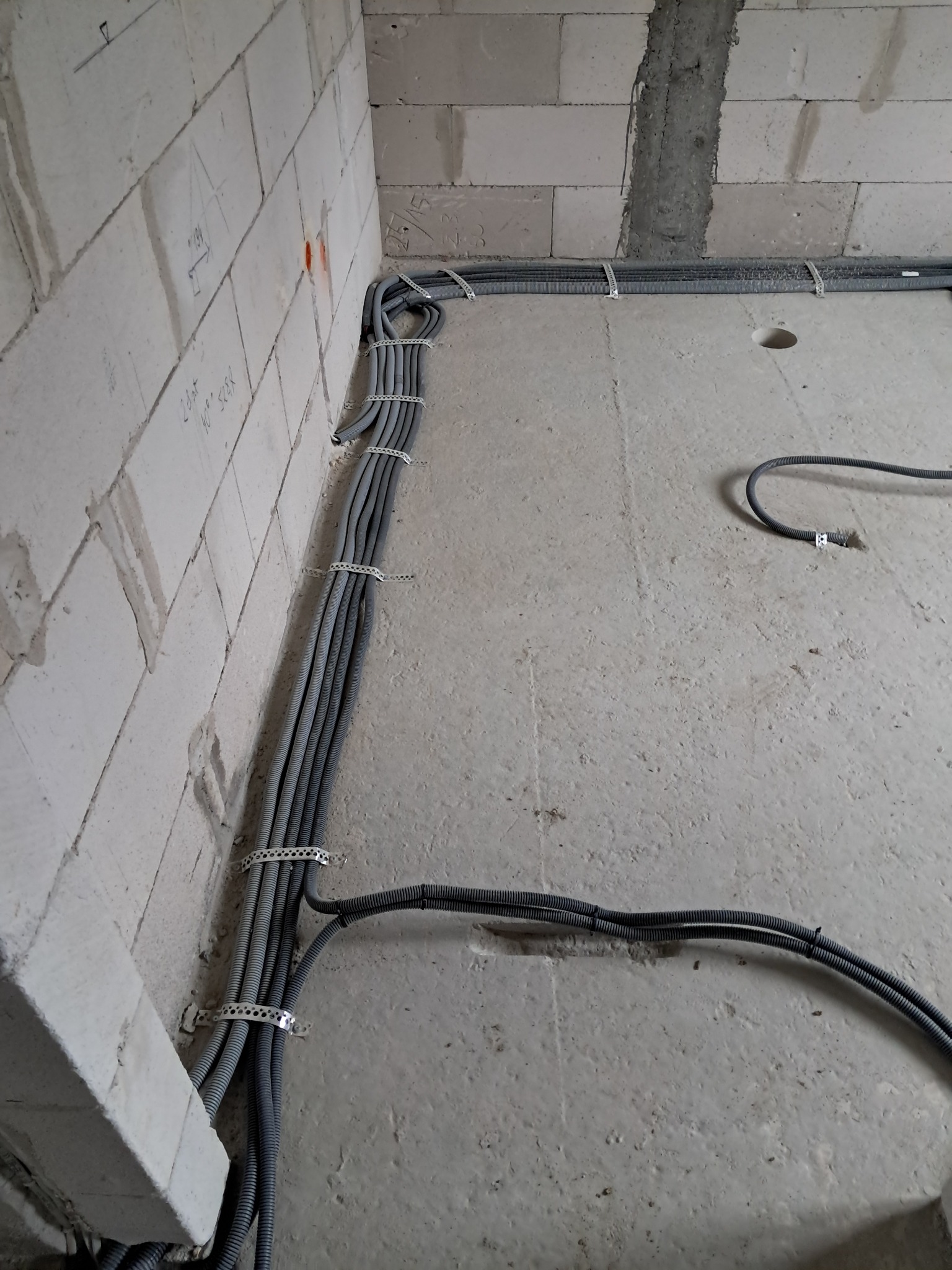 Szare rury instalacji elektrycznej przymocowane do ściany z białych bloczków i ułożone na szarej podłodze w surowym wnętrzu, widok z góry.