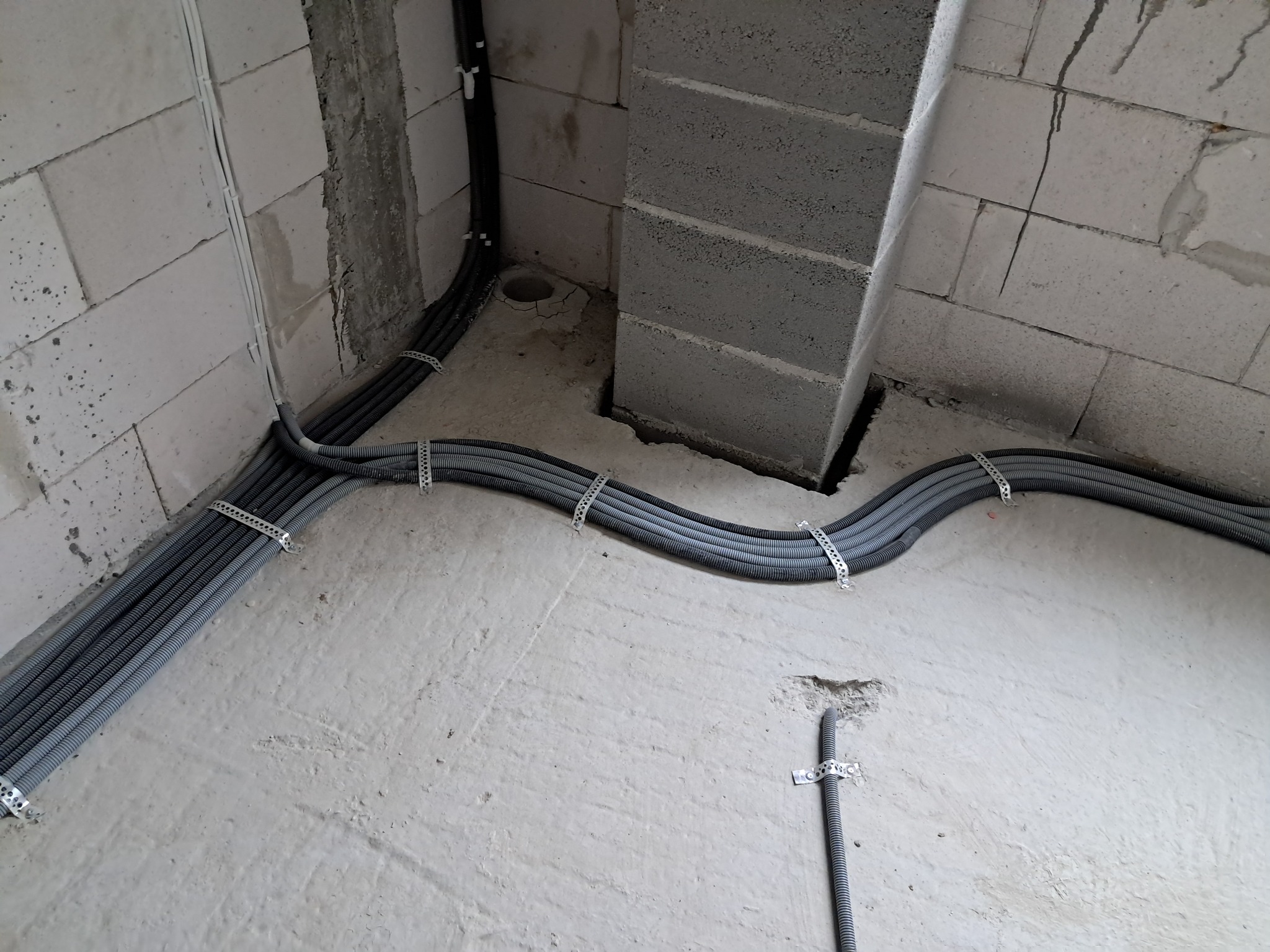 Szare rury instalacyjne, zabezpieczone metalowymi uchwytami, ułożone równolegle na surowej posadzce w budynku z białych bloczków. Widoczne otwory w podłodze i ścianach.