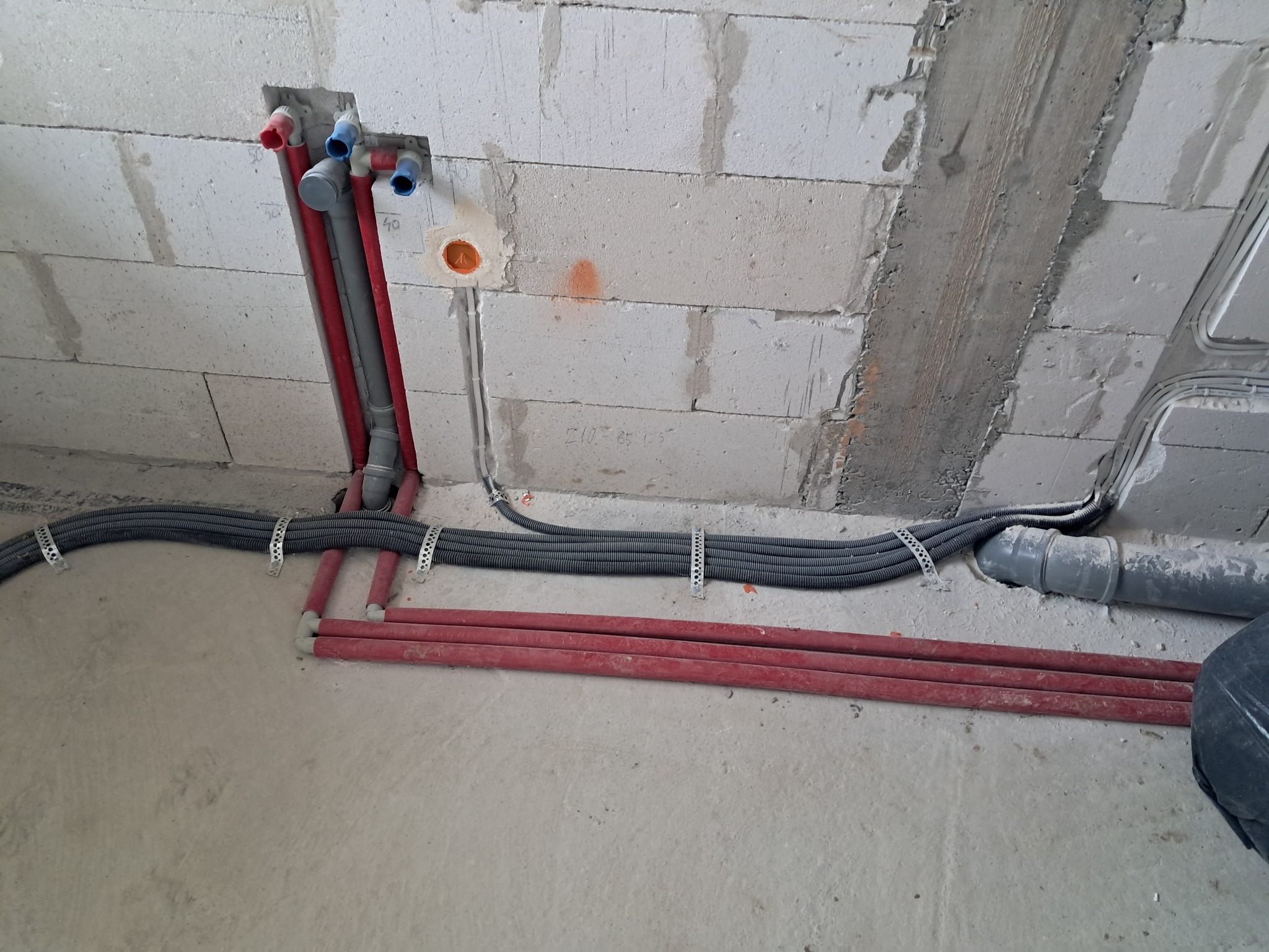 Surowy stan: Instalacja rur i przewodów w ścianie z betonu komórkowego. Widoczne peszle z kablami, rury wodne w kolorze czerwonym i szarym, puszka elektryczna w ścianie.